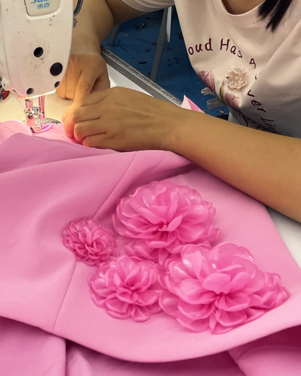 A machinist's hands guiding pink fabric under the machine foot, with five pink three-dimensional fabric flowers on the panel.