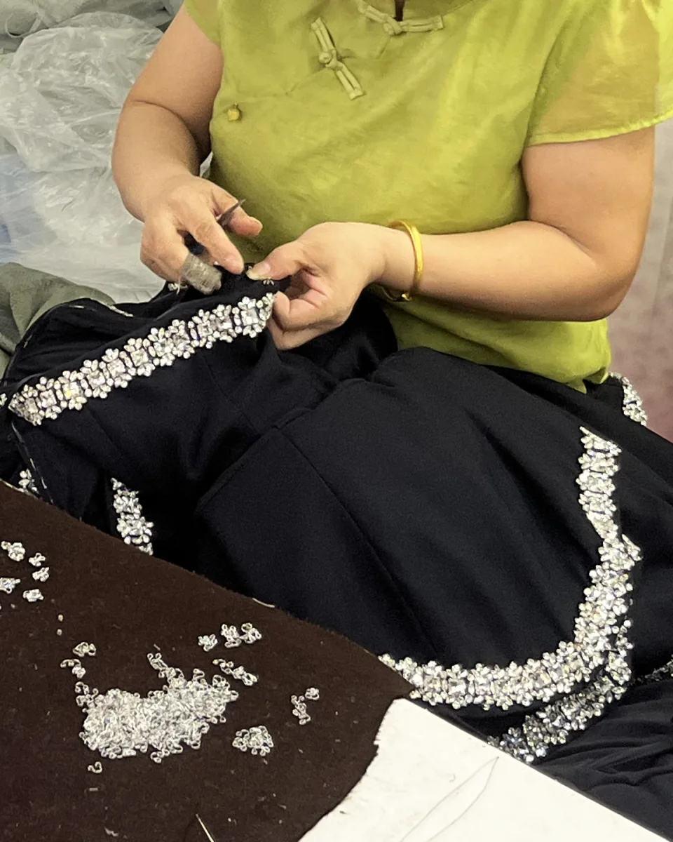 Hands working a band of clear stones along the edge of a black garment, with loose stones scattered on the table.