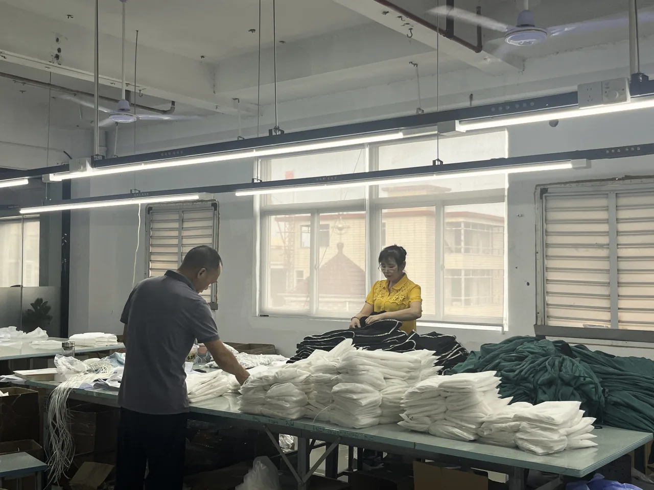 Two workers at a long table stacked with folded white garments.