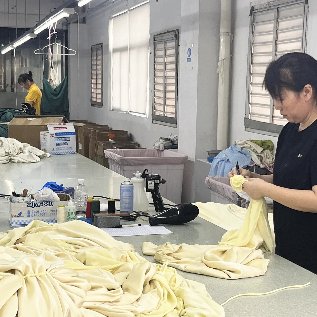 A worker trimming a pale yellow fabric flower by hand at the finishing table, more of the same fabric heaped in front.