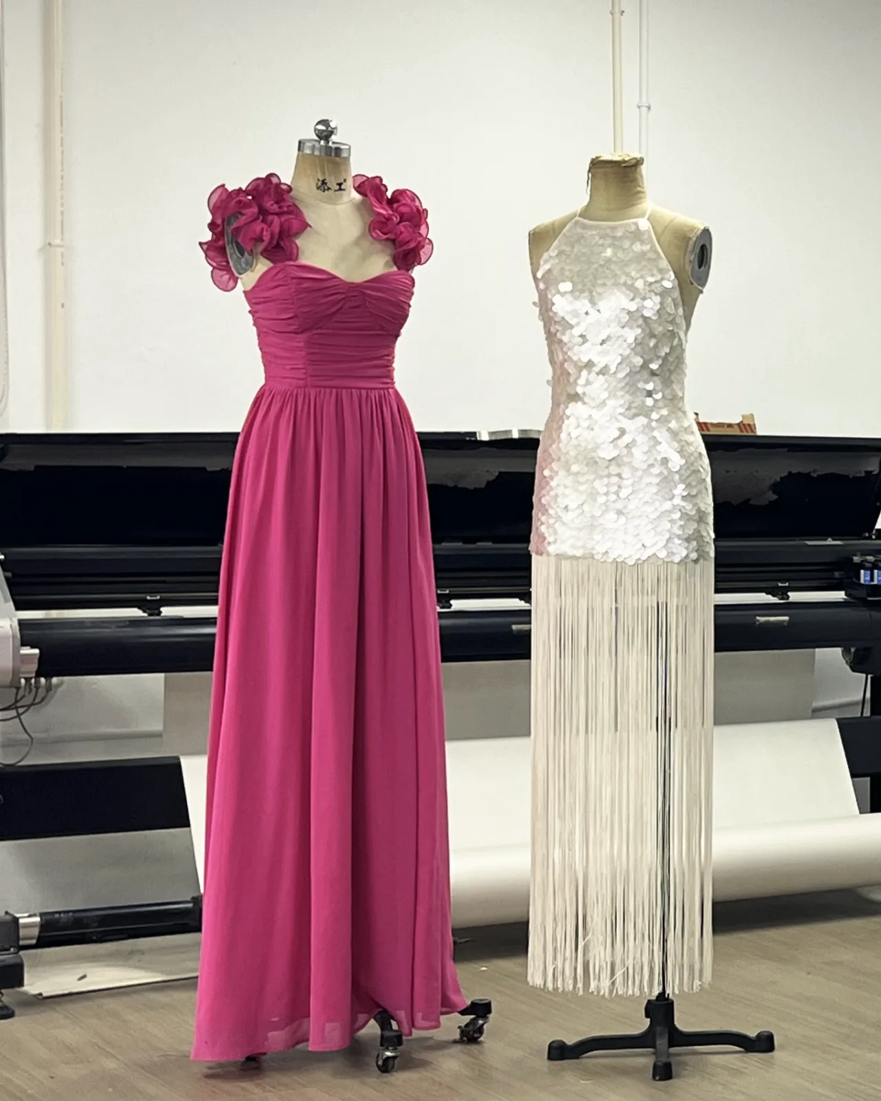 Two gowns on dress forms: a long pink chiffon gown with ruffled rosette shoulders, and a white paillette halter dress with a long fringe skirt.