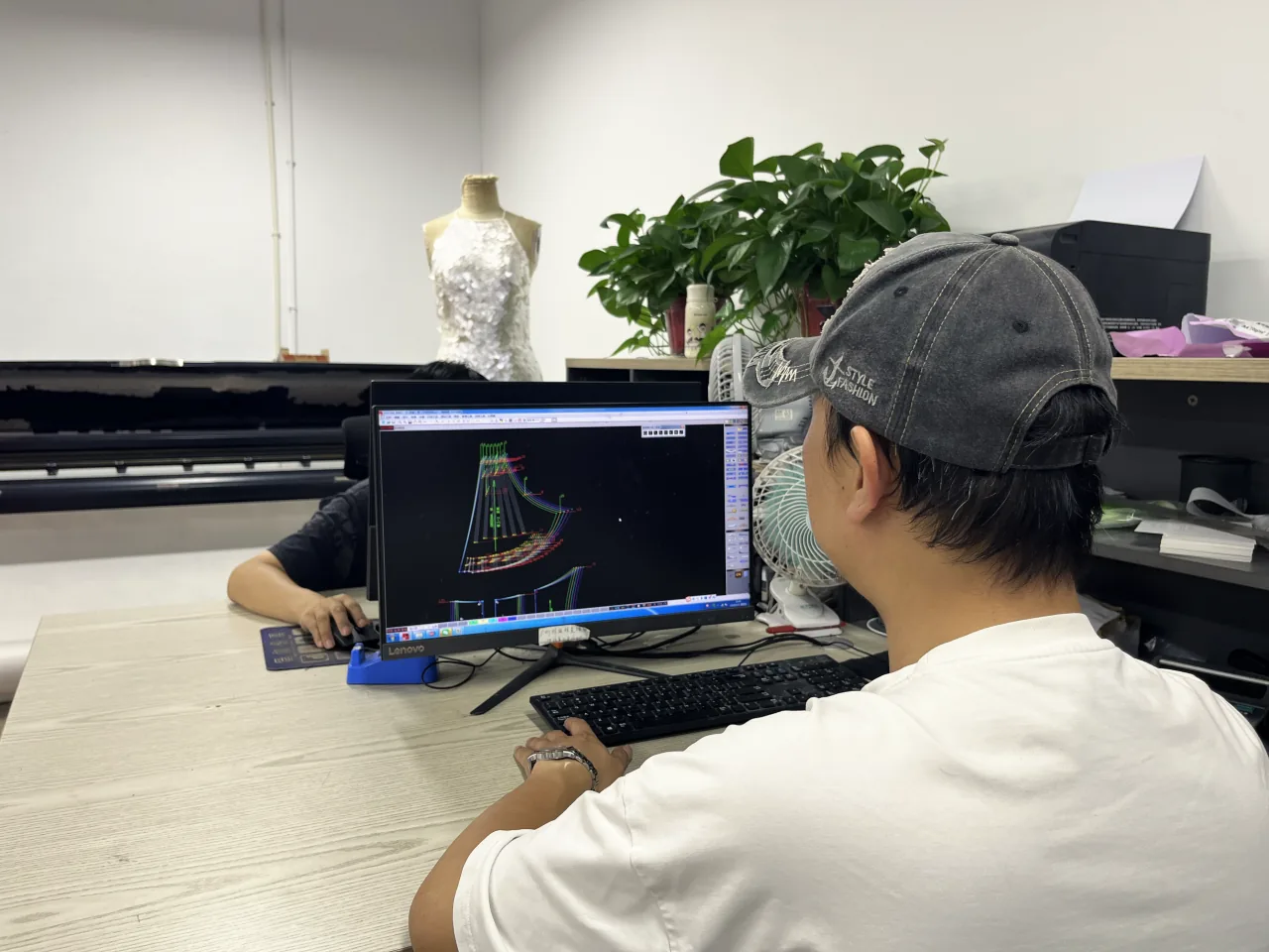 A pattern maker at a CAD screen, a sequinned sample on a dress form behind.