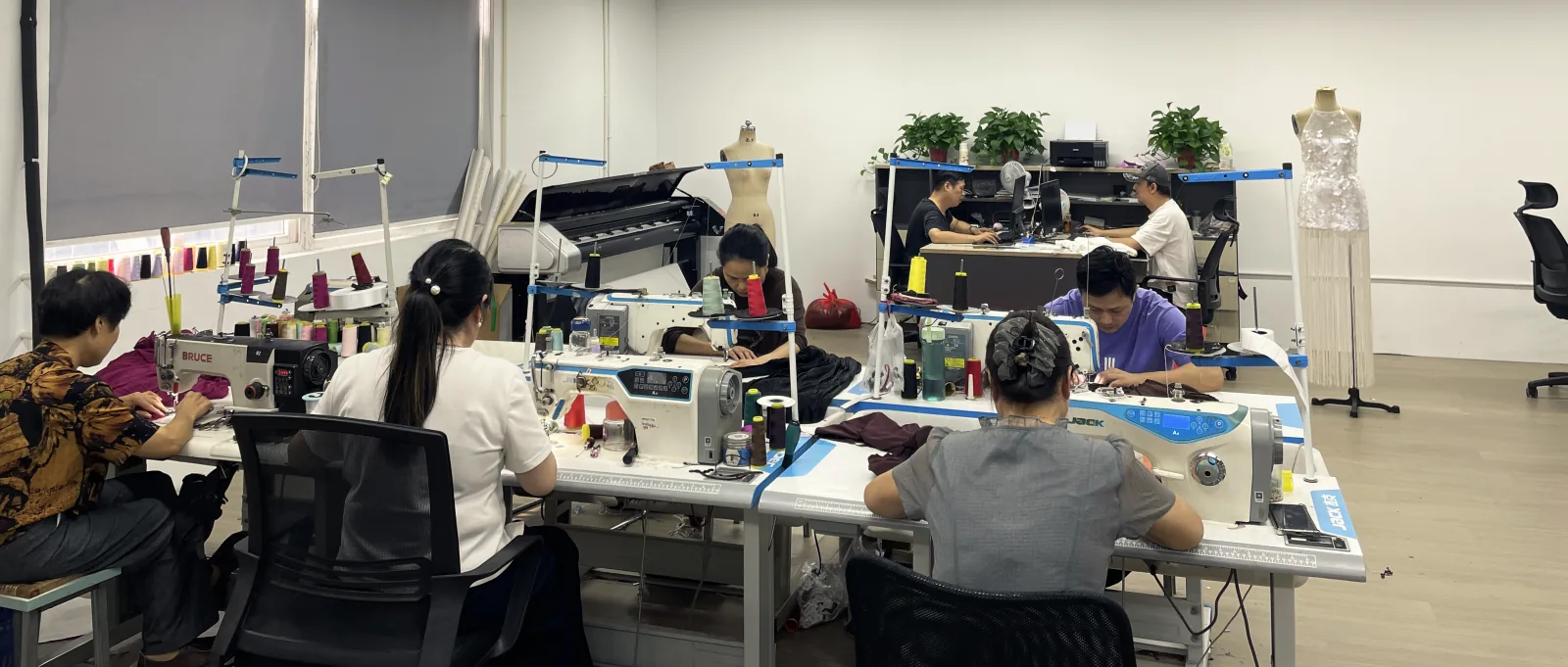 The sample room from the window side: machinists at a row of sewing machines, the pattern desk behind them, and a sequinned fringe dress on a dress form.