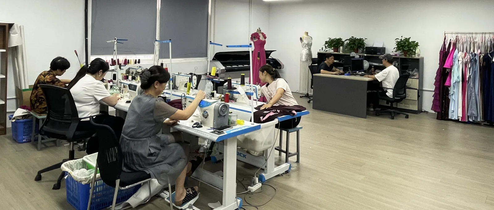 The sample room: machinists at their machines in front, a pink gown and a sequinned dress on dress forms, the pattern desk, and a rail of coloured dresses at the back.