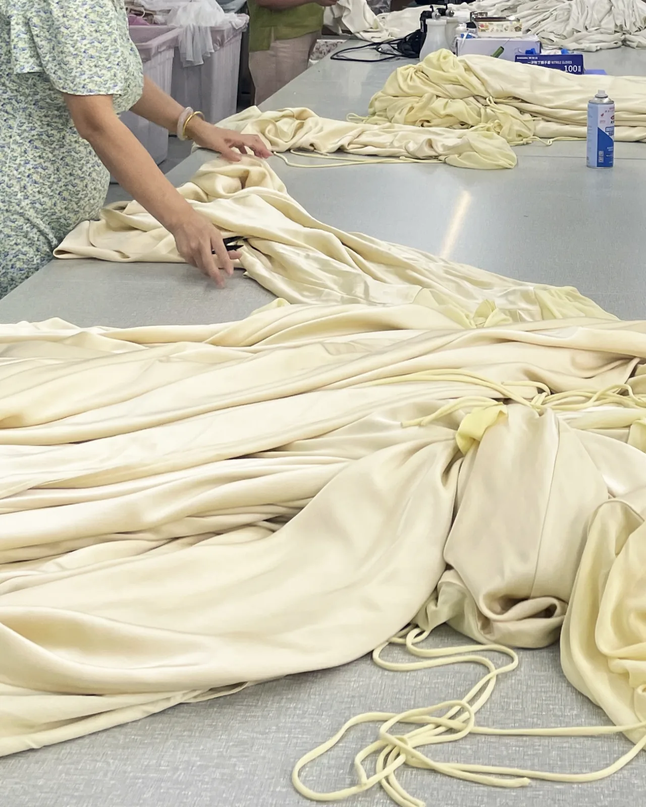 Hands smoothing cream satin-finish fabric pooled in folds across a work table, with loose drawcords.