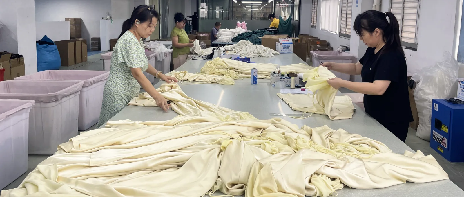A long finishing table heaped with cream satin-finish garments, two workers handling pieces in the foreground and more further down the room.