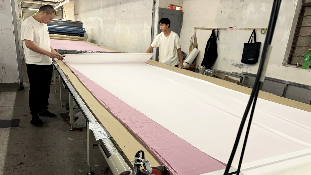 Two workers spreading layers of fabric along a long cutting table.